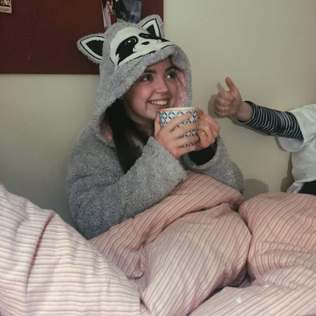 Girl sat in bed, wearing a onesie, and drinking a cup of tea