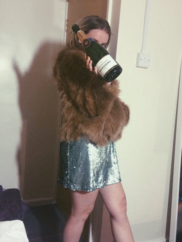 Girl wearing a glitter dress, fur coat, and posing behind a bottle of champagne