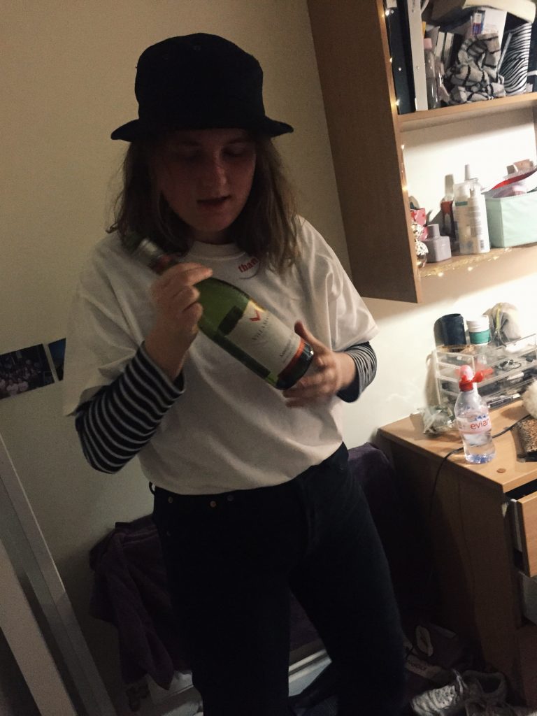 Guy with long hair, wearing a hat, posing with a bottle of wine