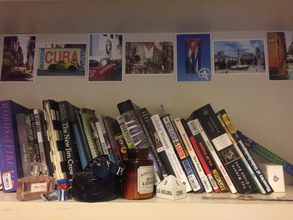 A bookshelf in my room decorated with Cuban postcards, the street sign replica, and a small drum keychain painted like the flag. An English version of "Memorias de Subdesarollo" sits at the end.