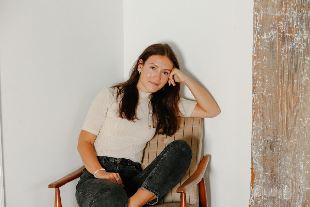 Emma Bates sat in a tartan chair, with legs crossed and her head casually resting on her hand. She wears black stonewashed jeans and a plain cream t-shirt.