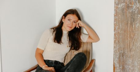 Emma Bates sat in a tartan chair, with legs crossed and her head casually resting on her hand. She wears black stonewashed jeans and a plain cream t-shirt.