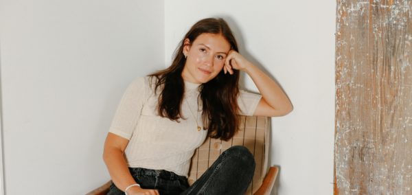 Emma Bates sat in a tartan chair, with legs crossed and her head casually resting on her hand. She wears black stonewashed jeans and a plain cream t-shirt.