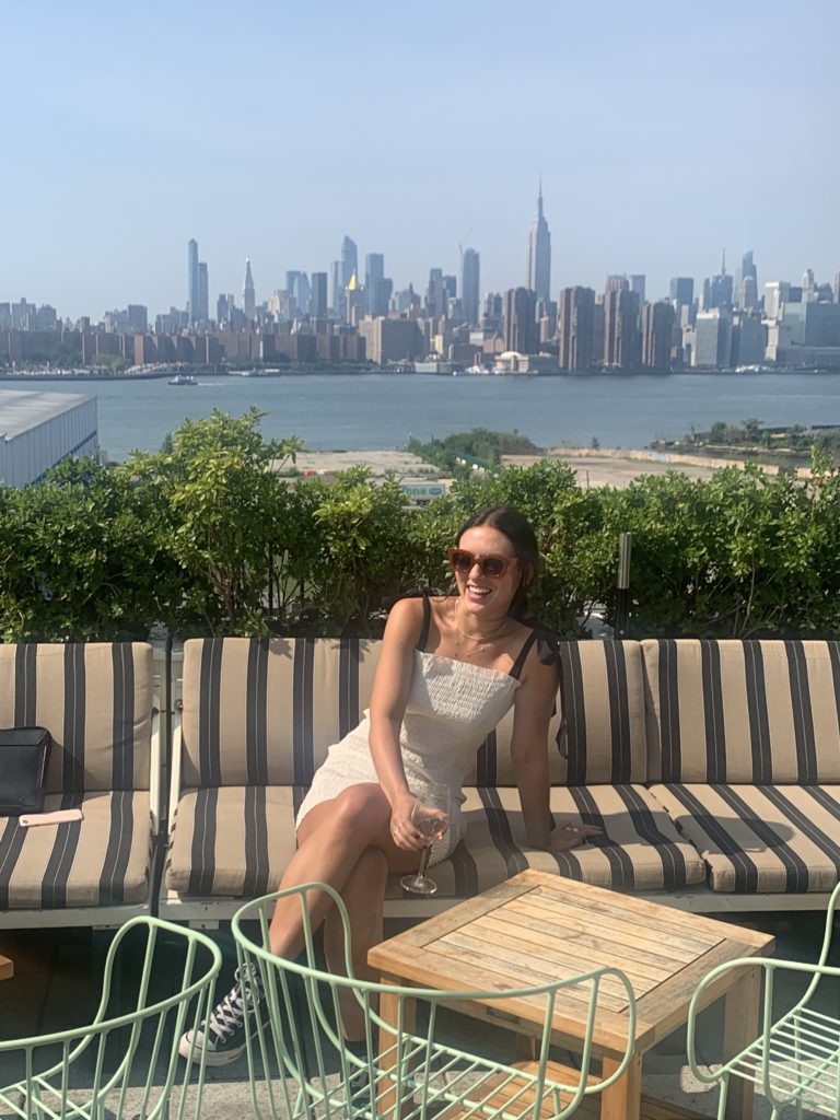 Emma Bates sits on the lounge bench of a sunny rooftop bar, wearing a white dress, retro sunglasses, and laughing as she looks off-camera. Behind her is the New York skyline.